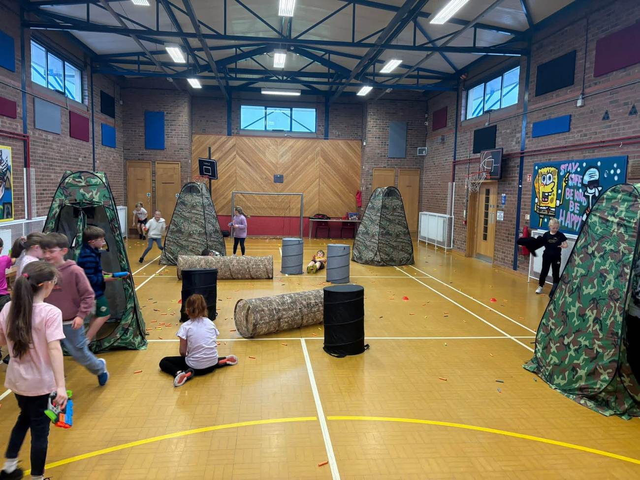 A full Nerf battle arena set up in a gym hall with tents, tunnels and barrels